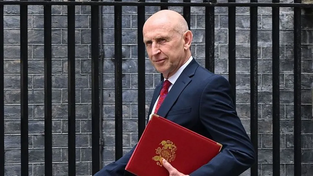 Getty Images Chancellor John Healey walks outside, wearing a navy suit with a red tie. He is holding a red folding and is looking to the side. Behind him is a brick wall and black railings.