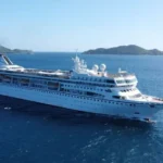 Villa Vie Residences A large white cruise ship sails through deep blue ocean waters near a chain of green, hilly islands under a clear sky.