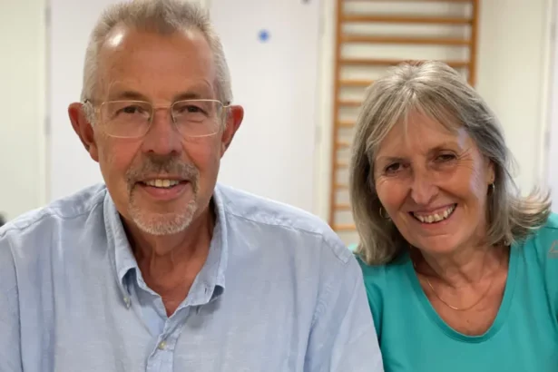 BBC Steve and Angela Bucksey sit side-by-side smiling - Steve has grey hair and is wearing a light blue shirt with a collar, and Angela has shoulder-length greyish hair and wears a green round-necked T-shirt and a necklace