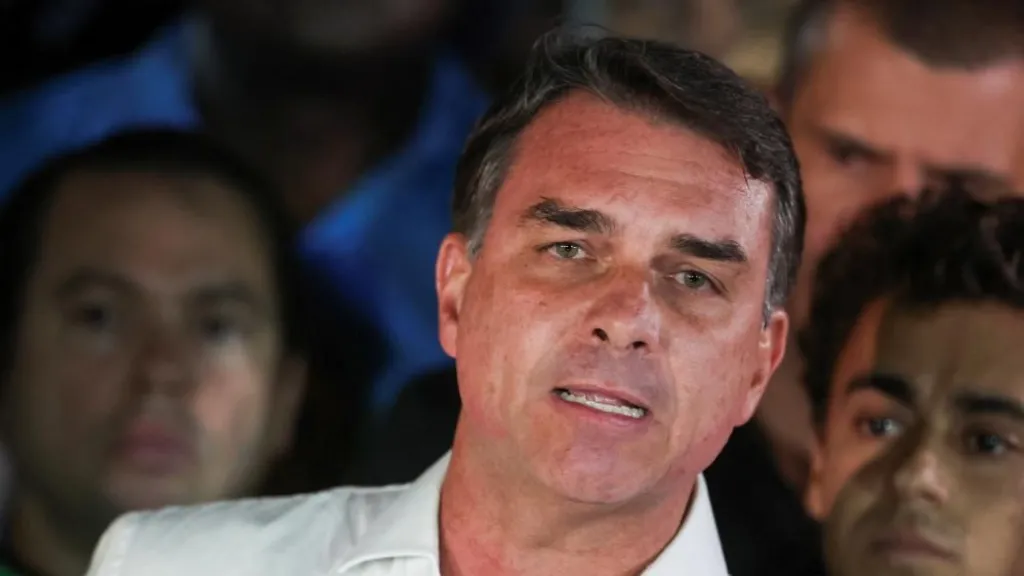 REUTERS/Adriano Machado Brazilian presidential candidate Flavio Bolsonaro of the Liberal Party speaks after losing the presidential runoff election, in Brasilia on 25 October, 2026. He is wearing a white shirt with an open collar. He is gesturing with one hand. Behind him, his supporters look on.