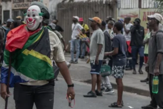 Getty Images A masked man draped in a South African flag stands in the street as onlookers gather behind him, holding a slingshot in one hand and a stick in the other.r
