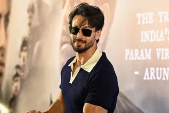 AFP via Getty Images Tiger Shroff poses at a promotional event, wearing dark sunglasses and a navy polo shirt with a white collar. The actor leans on a display stand in front of a backdrop featuring large portrait images and text from a film campaign.