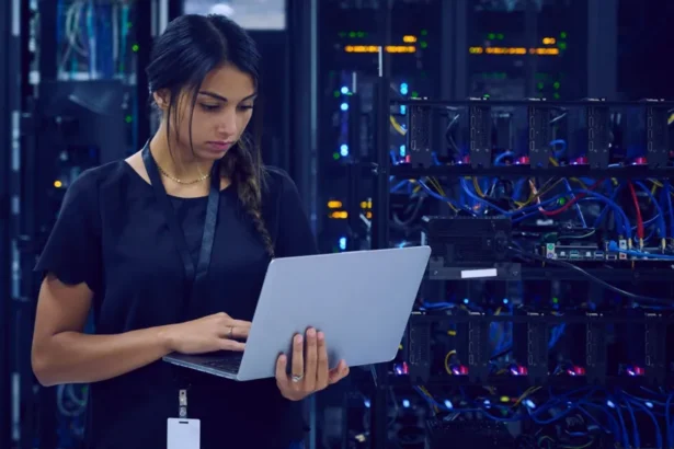 Getty Images A female technician dressed in navy blue clothes and wearing a lanyard around her neck is standing in a server room using her laptop.