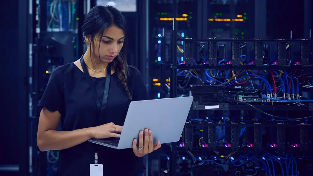 Getty Images A female technician dressed in navy blue clothes and wearing a lanyard around her neck is standing in a server room using her laptop.