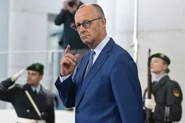 Getty Images German Chancellor Friedrich Merz in a blue suit with hand raised, two military officers stand behind him, blurred.