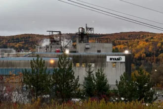 BBC/Jessica Murphy An image showing a part of the RYAM plant in Temsicaming, surrounded by red and orange fall trees. The building is grey with a sign in right corner that features the RYAM logo. Smoke is billowing out the ceiling.