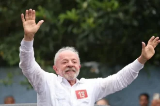 EPA/Shutterstock Luiz Inacio Lula da Silva greets supporters after casting his ballot. He is smiling and waving as he stands through the sunroof of a car.