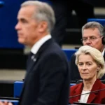 AFP via Getty Images An image showing European Commission President Ursula von der Leyen looking at an out of focus Mark Carney as he delivers a speech to the European Parliament. She has short blonde hair and is wearing a maroon jacket.