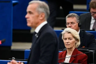 AFP via Getty Images An image showing European Commission President Ursula von der Leyen looking at an out of focus Mark Carney as he delivers a speech to the European Parliament. She has short blonde hair and is wearing a maroon jacket.