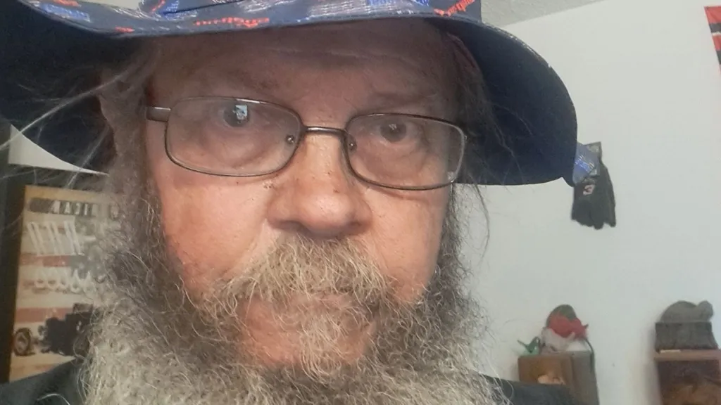 Geno Stecks Geno Stecks, 66, stares through eyeglasses at the camera, sporting a bushy beard, mustache and goatee under a floppy red-white-and-blue hat, a poster for an antique car visible in the background