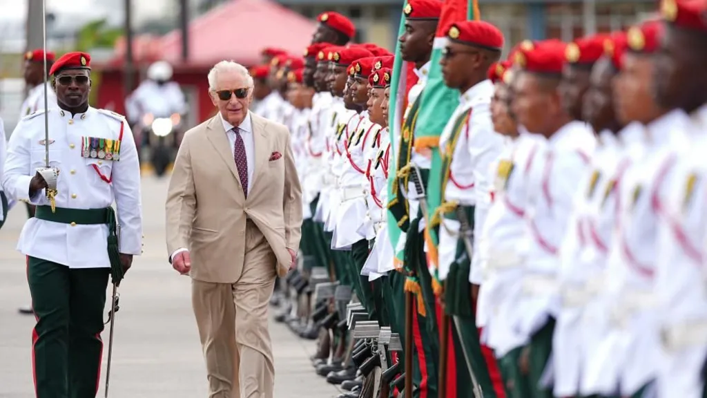 PA Media King Charles III, in a light brown suit, observes a guard of honour. He walks along a line of members of the Guyana Defence Force, who wear white shorts with military honours on them, and green trousers.