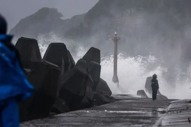 AFP via Getty Images Large waves crash against a coastal breakwater during stormy weather, sending spray high into the air. Massive concrete tetrapods line the left side of the seawall, while two people wearing blue rain gear stand on the wet waterfront.