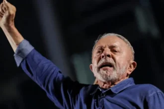 Reuters President Lula extends his right arm, his hand in a fist, as he reacts to his election. He is wearing a blue denim shirt, and has short grey hair and beard.
