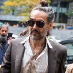 PA Russell Brand arrives at Southwark Crown Court, alongside his wife, wearing sunglasses and with his hair tied up, as cameras fix upon him