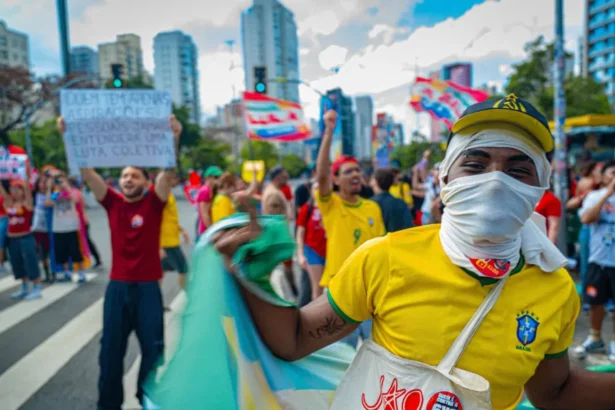 BBC A young man covering his face in a white cloth wearing a yellow Brazil football shirt waving a Brazil flag. In the Background protesters hold up flags and signs.