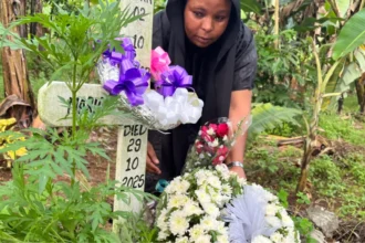 BBC Neema Munuo attending a grave with a simple cross and the words 'Died 29 10 2025' written on it. There are flowers and ribbons on the cross in a densely vegetated area