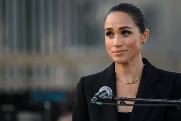 Getty Images Meghan Markle stands behind a microphone wearing a simple charcoal jacket and top, a gold necklace hangs across her chest. Her dark hair is pulled back in a bun.