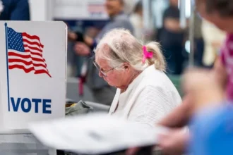EPA An older woman, wearing metal rimmed glasses, a white sweater, and with her white hair pulled up in a pink claw clip, sits in front of a voting booth. On it is a US flag and the word 'VOTE' in blue.