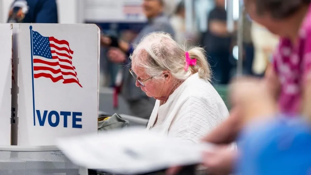 EPA An older woman, wearing metal rimmed glasses, a white sweater, and with her white hair pulled up in a pink claw clip, sits in front of a voting booth. On it is a US flag and the word 'VOTE' in blue.
