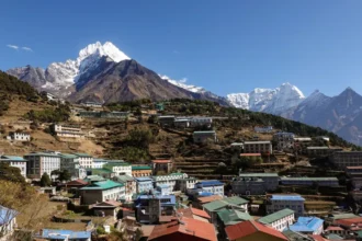 Didier Marti Namche Bazaar's colourful rooftops spread across terraced hillsides in Nepal's Everest region, with the snow-capped peak of Mount Thamserku towering above the town under a clear blue sky.