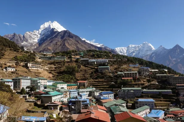 Didier Marti Namche Bazaar's colourful rooftops spread across terraced hillsides in Nepal's Everest region, with the snow-capped peak of Mount Thamserku towering above the town under a clear blue sky.