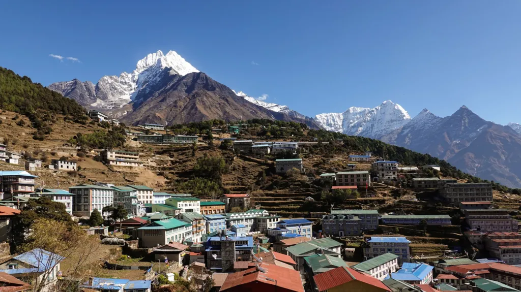 Didier Marti Namche Bazaar's colourful rooftops spread across terraced hillsides in Nepal's Everest region, with the snow-capped peak of Mount Thamserku towering above the town under a clear blue sky.