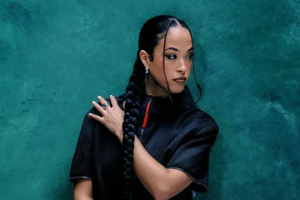 Supplied Jazzy is a 30-year-old woman with long dark hair which she wears in a braid down to her middle, with two smaller braids shaping her face. She looks over her left shoulder, her left arm across her body. She's photographed wearing a dark short sleeved shirt with a red zip against a blue backdrop.
