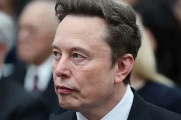 Getty Images Elon Musk looks sideways as he wears a dark suit, white shirt and grey tie, with other people visible in the background.