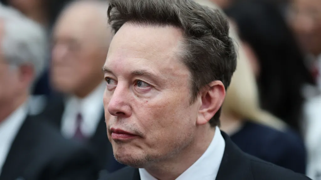 c94ca0d0-d041-11f1-950a-2dcbf5245e34-jpg Getty Images Elon Musk looks sideways as he wears a dark suit, white shirt and grey tie, with other people visible in the background.