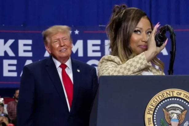 Reuters Nikki Minaj at the microphone at a podium with the presidential seal. She has a slight smile and a gold suit with a Gucci logo. Trump smiles behind her.