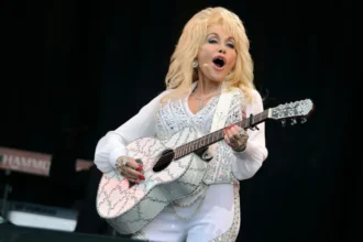 BBC Dolly Parton is holding an acoustic guitar and wearing a white outfit while singing on stage.