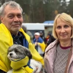 BBC Viacheslav, who has grey hair and is draped in a Ukrainian flag, holds a small dog in his arms. He is stood next to his wife Olha, who has a blonde bob. Both are looking at the camera.