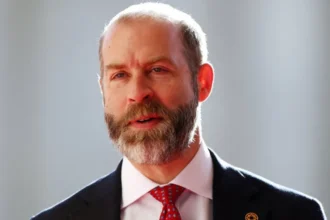 Reuters Head and shoulders shot of Reynolds, wearing a black jacket over a white shirt and red tie. The backdrop is out of focus and pale but sunny.