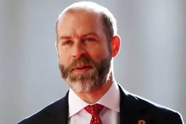 Reuters Head and shoulders shot of Reynolds, wearing a black jacket over a white shirt and red tie. The backdrop is out of focus and pale but sunny.