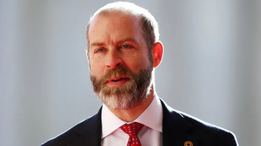 Reuters Head and shoulders shot of Reynolds, wearing a black jacket over a white shirt and red tie. The backdrop is out of focus and pale but sunny.