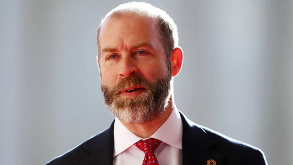 Reuters Head and shoulders shot of Reynolds, wearing a black jacket over a white shirt and red tie. The backdrop is out of focus and pale but sunny.