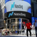 Michael Nagle/Bloomberg via Getty Images Two men walk in front of the Nasdaq building, which has a large curved lit up sign that reads Nasdaq on the front.