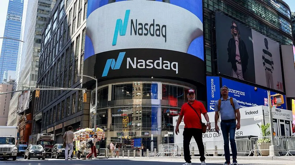 dcdf2a60-d46e-11f1-9dda-1ba6fcc7d159-jpg Michael Nagle/Bloomberg via Getty Images Two men walk in front of the Nasdaq building, which has a large curved lit up sign that reads Nasdaq on the front.