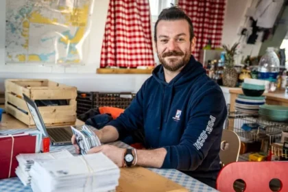 UK Antarctic Heritage Trust George Clarke sitting at a table with a stack of letters. He is wearing a hoodie with Port Lockroy written on the arm. A kitchen can be seen behind with a sash window.