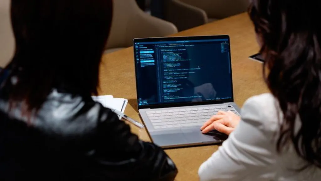 eba975d0-cdcc-11f1-bd0d-75577ccf219c-jpg Getty Images The image shows two people from behind as they sit at a table looking at code on a laptop.