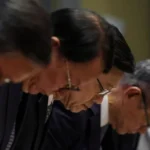 Reuters A row of Chinese men wearing dark suits and glasses bow in apology, with the focus of the shot centering on John Lee in the middle of the row