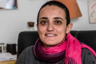 AFP via Getty Images Egyptian journalist Lina Attalah sits on a black leather chair. She has a red scarf around her neck, a grey top, cropped black hair, stud earrings and and a silver metal studd in her right nostril, a broad smile on her lips.