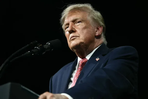 Kent NISHIMURA / AFP via Getty Images President Trump stands against a black background behing a black podium/ He is wearing a white shirt, blue suit coat with an American flag pin and a red print tie. He is not smiling and is gripping the edges of the podium. He is photographed from below the podium looking up.
