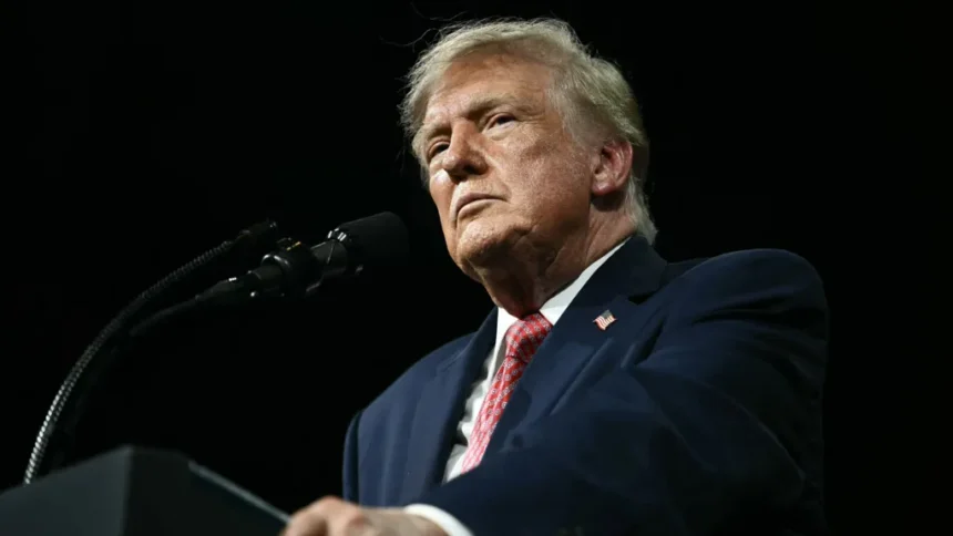 Kent NISHIMURA / AFP via Getty Images President Trump stands against a black background behing a black podium/ He is wearing a white shirt, blue suit coat with an American flag pin and a red print tie. He is not smiling and is gripping the edges of the podium. He is photographed from below the podium looking up.